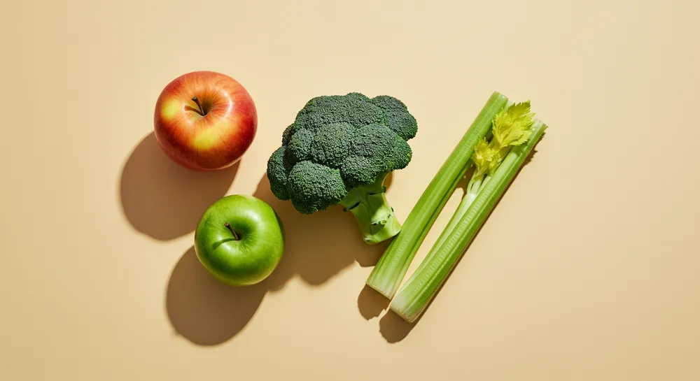 Fresh red apple, green apple, broccoli, and celery stalks on a beige surface.